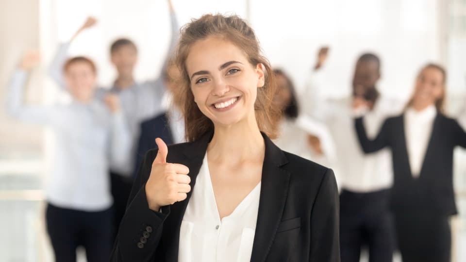A smiling HR manager gives a thumbs-up with a cheering business team in the background after a successful relocation process.