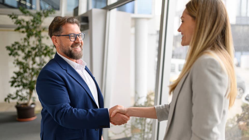 Successful B2B project completion at DACHSER & KOLB – A smiling project manager in a blue suit shakes hands with a client in a modern office.