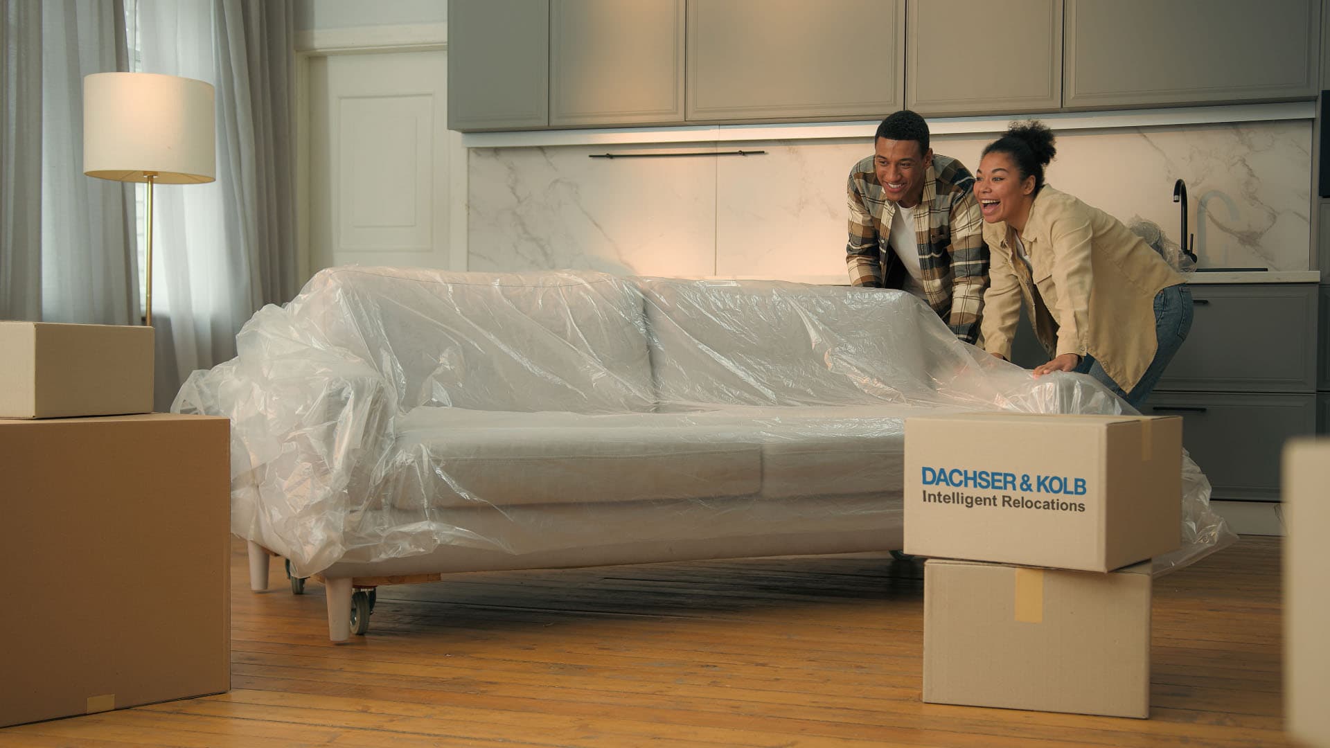 Two people move a sofa wrapped in protective film inside an apartment among moving boxes