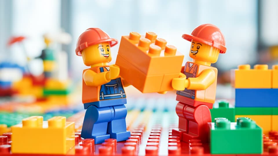 Zwei Minifiguren als Umzugshelfer mit Schutzhelmen transportieren gemeinsam einen großen Baustein – professioneller Umzugsservice von DACHSER & KOLB.