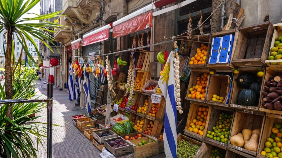 Straße mit Einkaufsläden in Uruguay, vor denen Kisten mit Obst und Gemüse aufgebaut sind