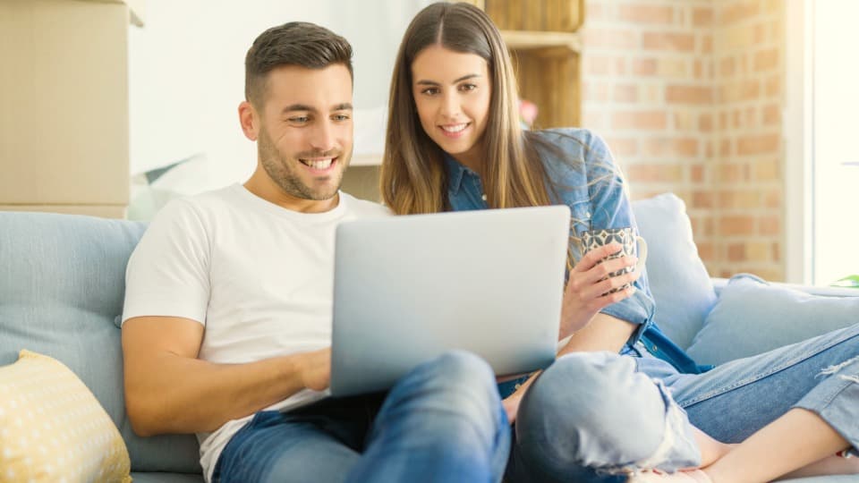 A happy couple relaxes on a couch in front of moving boxes, using a laptop to finance their move with DACHSER & KOLB via installment payments.