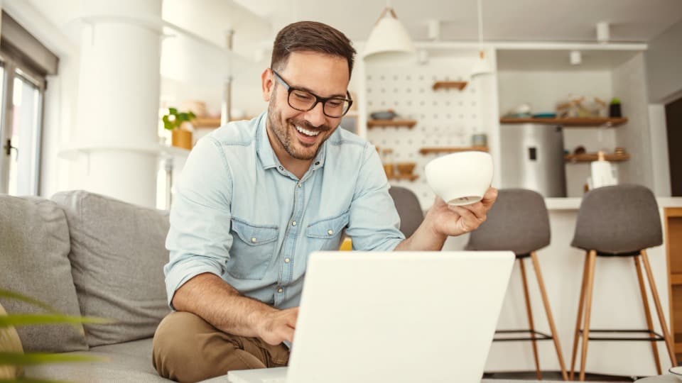 A young man sits laughing on a couch with a coffee cup, using the DACHSER & KOLB online calculator to get his moving quote.