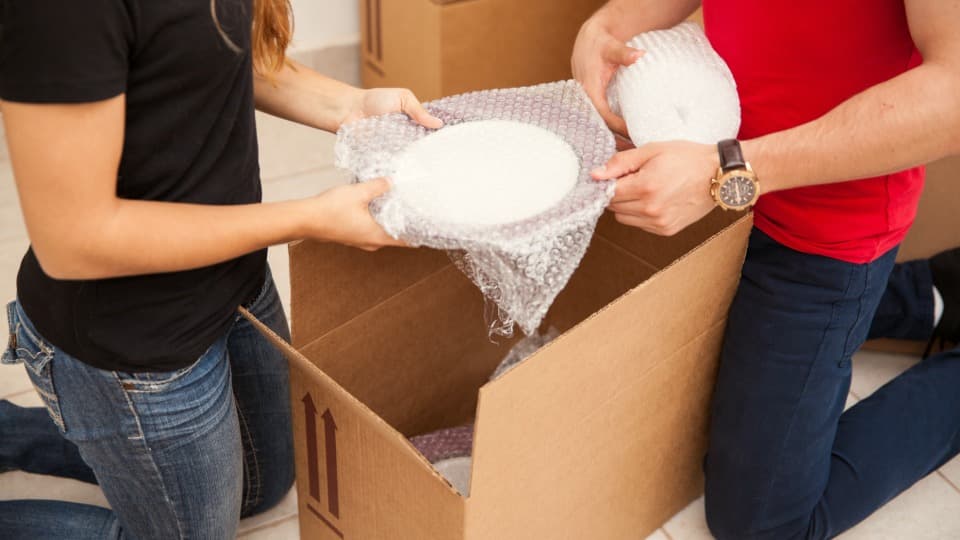 Two people wrapping fragile dishes in bubble wrap and packing them into a moving box in preparation for transport with DACHSER & KOLB.