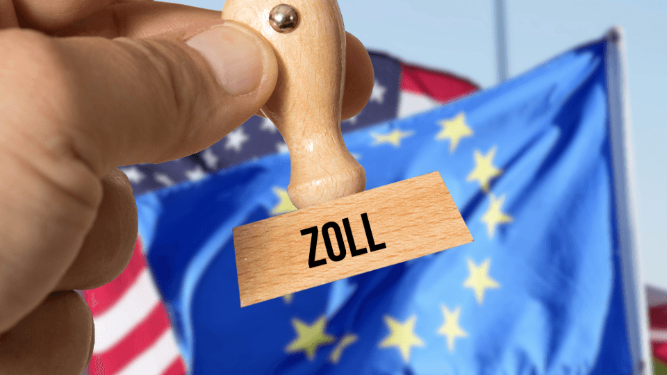 Hand holding a wooden stamp with the word ZOLL in front of USA and EU flags