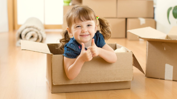 A happy little girl giving a double thumbs up from inside a moving box during a relaxed international family relocation with DACHSER & KOLB.