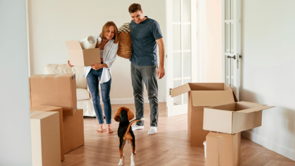 Ein glückliches Paar trägt gepackte Umzugskartons in einer hellen Wohnung als Vorbereitung für den Kleintransport mit DACHSER & KOLB.
