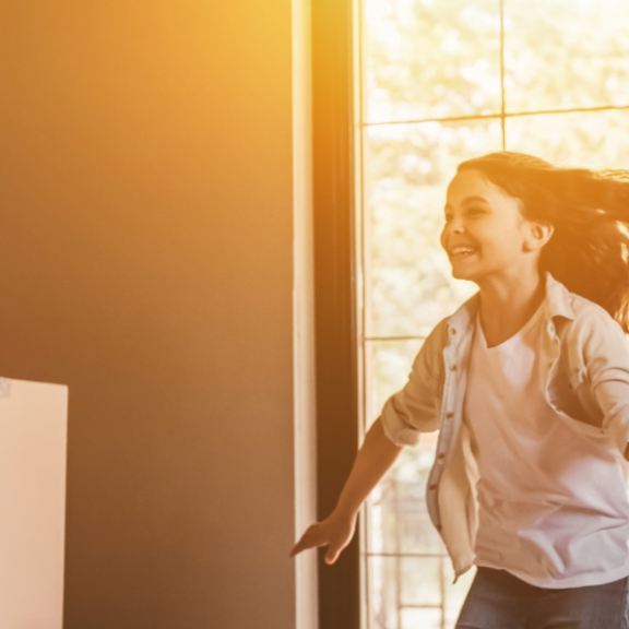 Fröhliche Familie beim Privatumzug – Kind läuft lachend ins neue Zuhause, Eltern tragen Umzugskartons ins Haus.