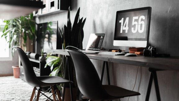 Graues Home Office nach Umzug mit langem Schreibtisch, zwei Stühlen, Laptop, Monitor mit Uhrzeit und Pflanzen. Einrichtungsideen für gemeinsames Arbeiten.