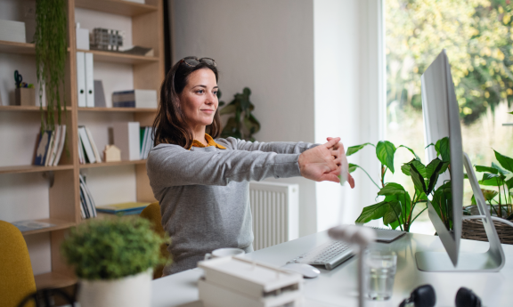 Frau dehnt Handgelenke im hellen Home Office nach Umzug mit Schreibtisch, Computer und Pflanzen. Einrichtung für gesundes Arbeiten zu Hause.