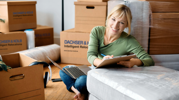 Frau plant den Umzug mit Klemmbrett und Laptop zwischen Umzugskartons