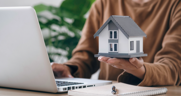 Frau prüft Versicherung nach dem Umzug online und hält ein Modellhaus als Symbol für Immobilienabsicherung.