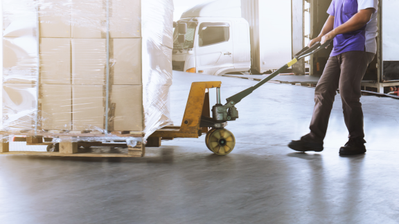 Arbeiter zieht einen Hubwagen mit verpackten Kartons. Keine Holzkisten (Crates) sichtbar. Lager und Logistik.