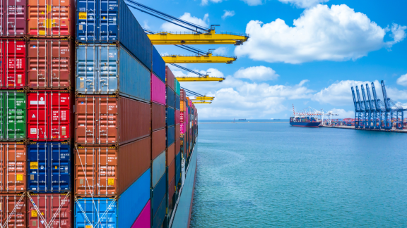 Ein großes Containerschiff beladen mit farbigen Containern verschiedener Größen im Hafen unter blauem Himmel.