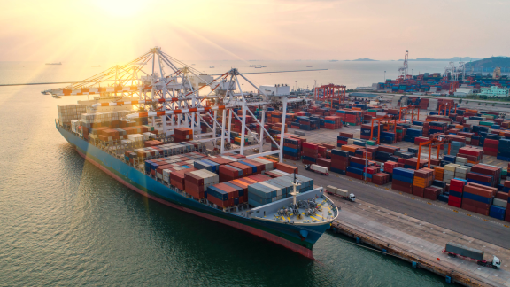 Ein großes Containerschiff im Hafen, beladen mit FCL-Containern. Kräne verladen Fracht in der Abendsonne.